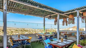 An outdoor dining area with tables and chairs, a pergola with hanging plants, and a view of a cityscape. - Cerro Colorado Condo Condo in Cabo San Lucas (Cabo San Lucas)