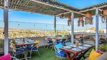 An outdoor dining area with tables and chairs, a pergola with hanging plants, and a view of a cityscape.