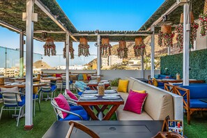 Un patio al aire libre con mesas y sillas, un sofá, cestas de flores colgantes y vistas del paisaje.