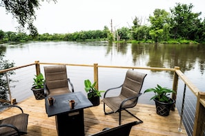 A wooden deck with two chairs and a table, surrounded by potted plants, overlooking a calm river. - Newly Remodeled Riverfront Cottage (Rockford)