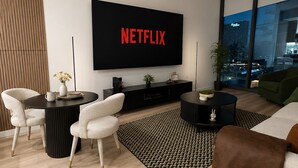 A modern living room with a large flat-screen TV displaying 'NETFLIX,' a round coffee table, two white armchairs, a sofa, and a view of a cityscape through large windows. - Prime Atlanta Location 2026 Flat close to everything! (Atlanta)