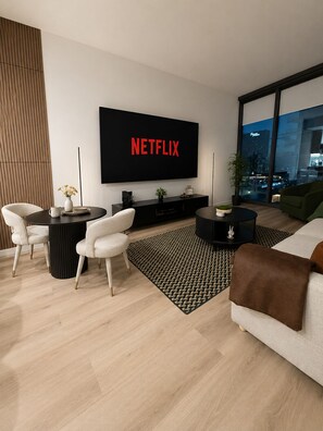 Ein modernes Wohnzimmer mit großem Flachbildfernseher, der 'NETFLIX' zeigt, einem runden Couchtisch, zwei weißen Sesseln, einer Couch und Blick auf eine Stadtlandschaft durch große Fenster.