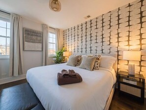 Habitación de hotel con una cama grande, una mesita de noche con lámpara, una pared con diseño geométrico y una ventana con cortinas.