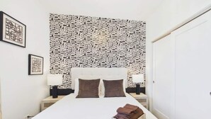 Een net opgemaakt bed met een hoofdbord met patroon, twee bedlampjes en een klein nachtkastje met een lamp.