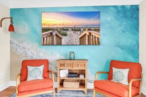 A room with two orange chairs, a wooden side table, and a beach-themed wall mural. - Oceanfront top-floor condo steps from the sand in North Myrtle Beach (North Myrtle Beach)