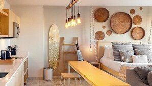 Espaço compacto com cozinha compacta, cama com várias almofadas, um banco de madeira e uma prancha de surf.