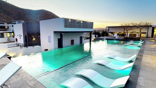 Piscina com espreguiçadeiras, um edifício moderno e vista para uma colina.