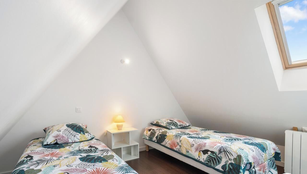 Una habitación pequeña y triangular con dos camas, una mesita de noche, una lámpara y una ventana.