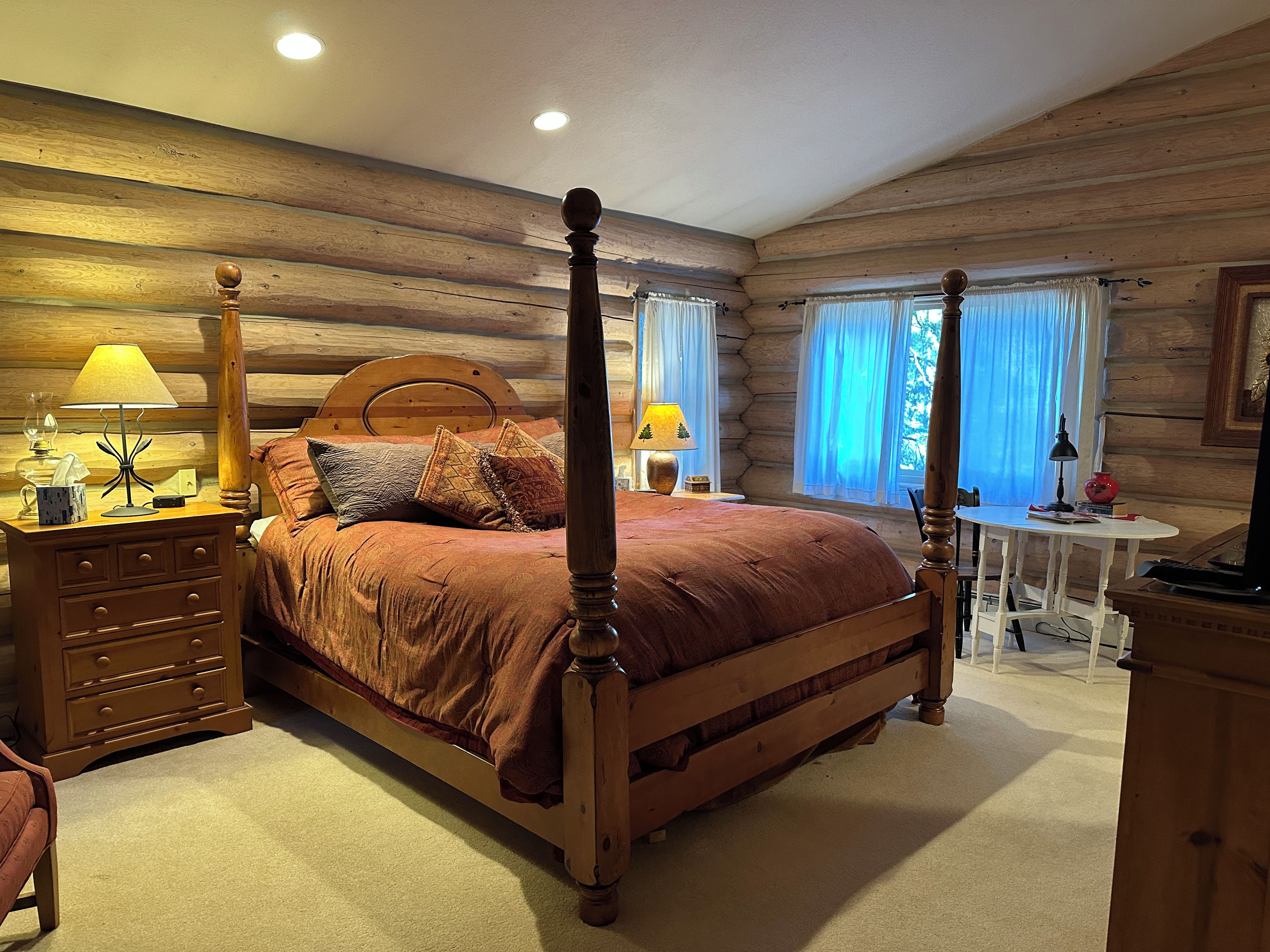 A log cabin bedroom with a four-poster bed, two bedside tables with lamps, a small round table with a chair, and a television.