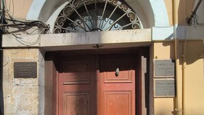 Een dubbele houten deur met een metalen hekwerk erboven, geplaatst in een gebouw met een stenen gevel en een klein rondboogvenster boven de deur.