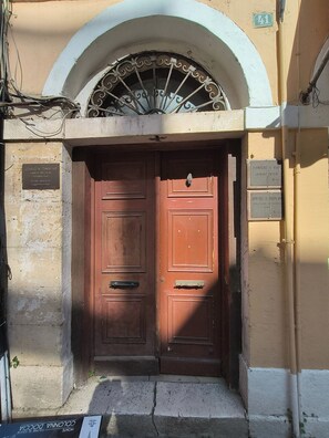 Eine doppelte Holztür mit einem Metallgitter darüber, in eine Fassade aus Stein eingelassen, oberhalb der Tür befindet sich ein kleines rundbogenförmiges Fenster.
