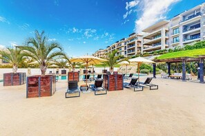 A resort with outdoor seating, palm trees, a pool, and modern buildings in the background. - Ground-floor Condo With Ocean View, Pool, Gym, Hot Tub, & Washer/dryer (Cabo San Lucas)