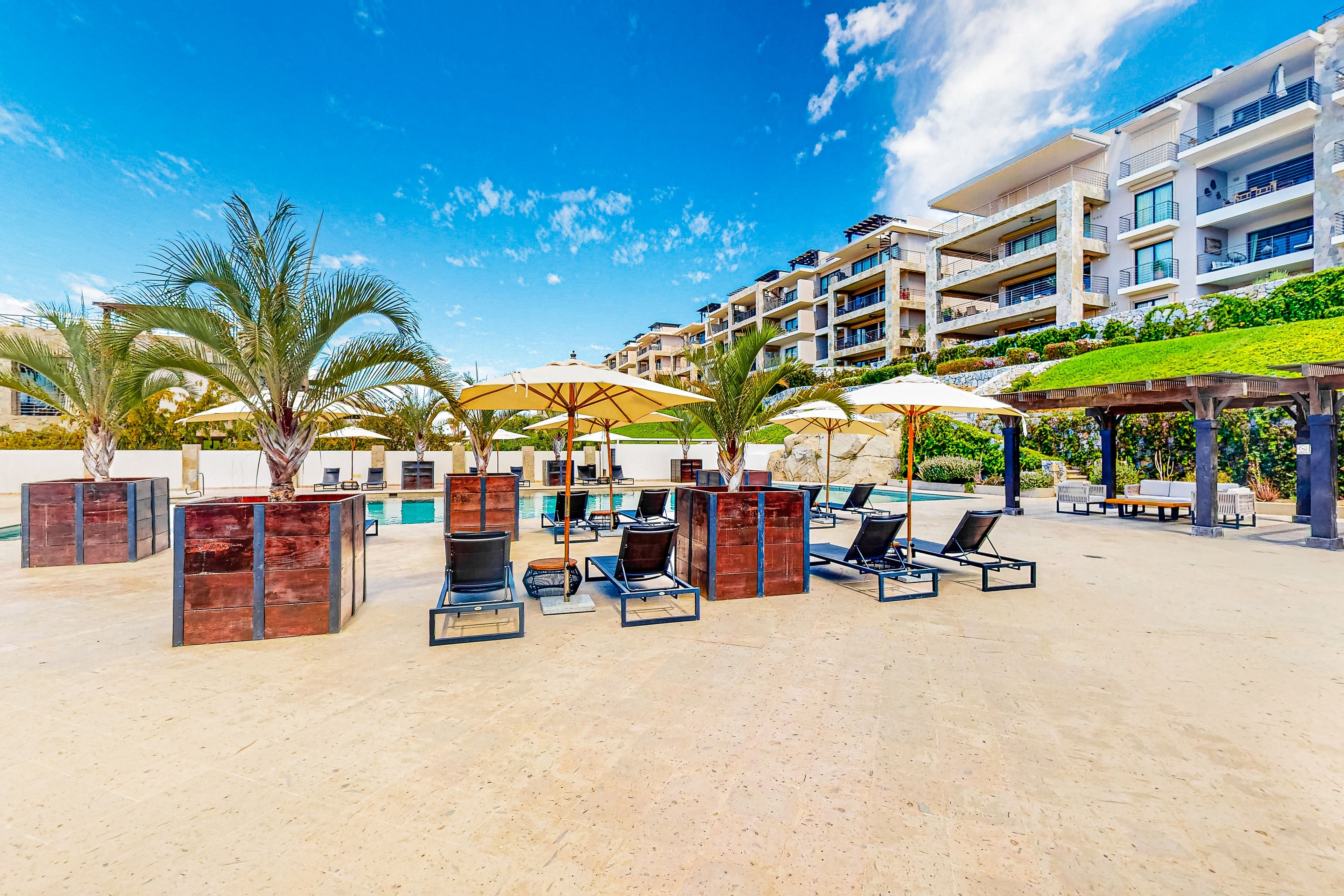 A resort pool area with sun loungers, umbrellas, and palm trees.