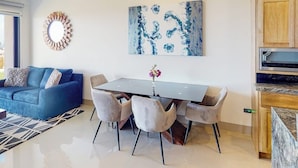 Een moderne woonkamer met een blauwe bank, een eettafel met stoelen, een abstract schilderij aan de muur en een keuken met houten kastjes.