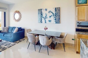 A modern living room with a blue sofa, a dining table with chairs, a wall-mounted abstract painting, and a kitchen area with wooden cabinets. - Ground-floor Condo With Ocean View, Pool, Gym, Hot Tub, & Washer/dryer (Cabo San Lucas)