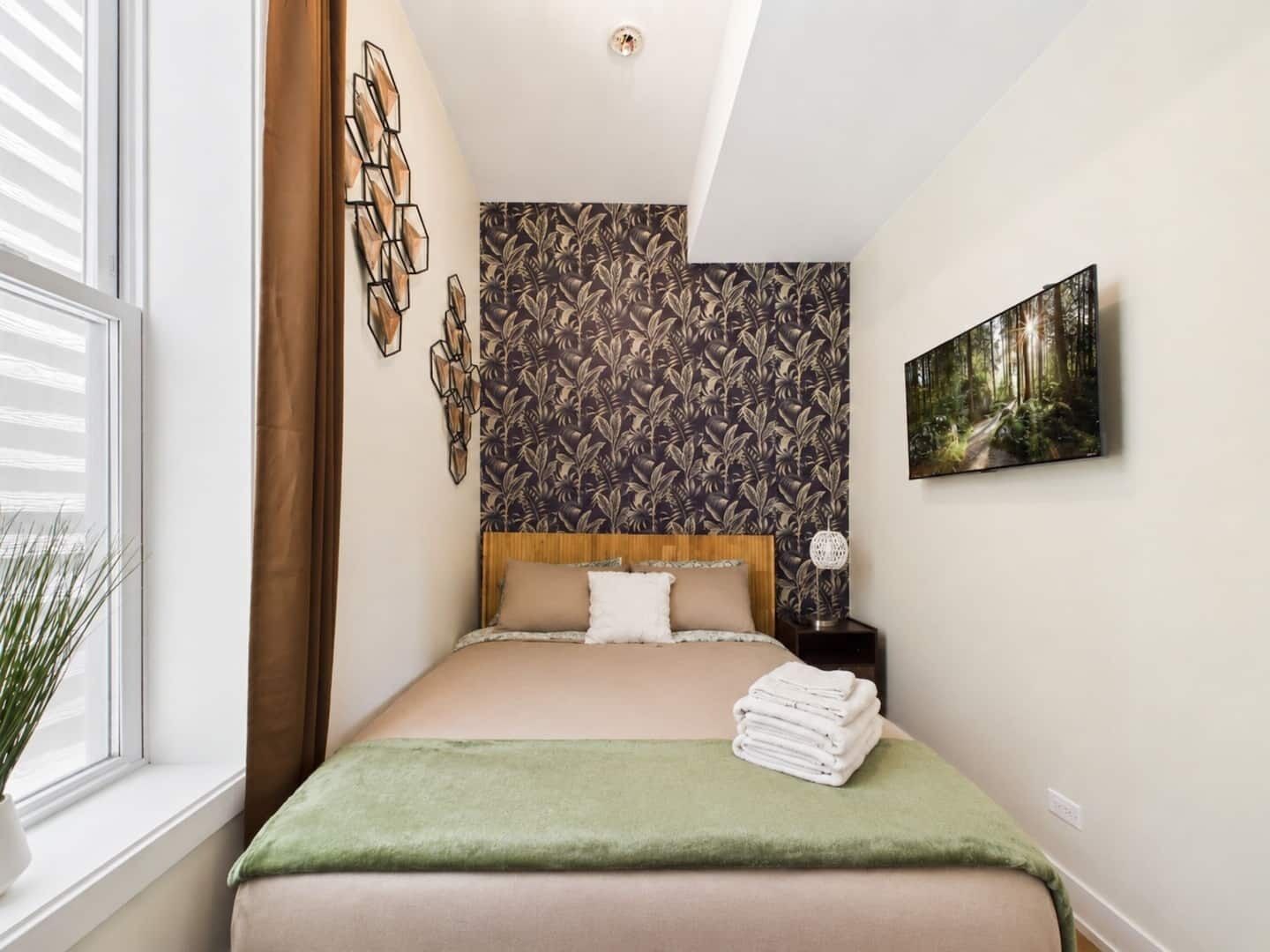 A bedroom with a bed, a wall with a leaf pattern, a painting, and a window with blinds.