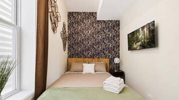 A bedroom with a bed, a wall with a leaf pattern, a painting, and a window with blinds.