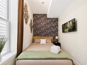 Een slaapkamer met een bed, een muur met bladpatroon, een schilderij en een raam met zonwering.