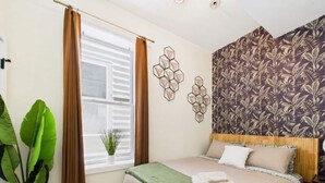 A bedroom with a bed, a window with curtains, a wall with a geometric pattern, and a potted plant.