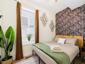 Une chambre à coucher avec un lit, une fenêtre avec des rideaux, un mur orné d’un motif géométrique et une plante en pot.