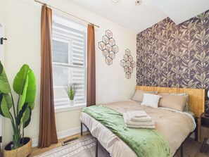 Quarto com cama, janela com cortinas, parede com padrão geométrico e uma planta grande.