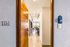 Un couloir avec une porte en bois portant le numéro « 104 », une boîte aux lettres bleue et une vue sur un espace extérieur aménagé avec des sièges.