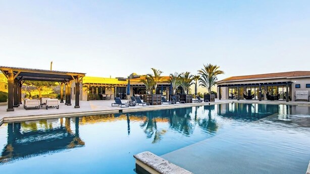 A swimming pool with lounge chairs and a covered area, surrounded by palm trees. - Modern Apartment With Ocean View, Pool, Patio, Tennis & Gym (Cabo San Lucas)