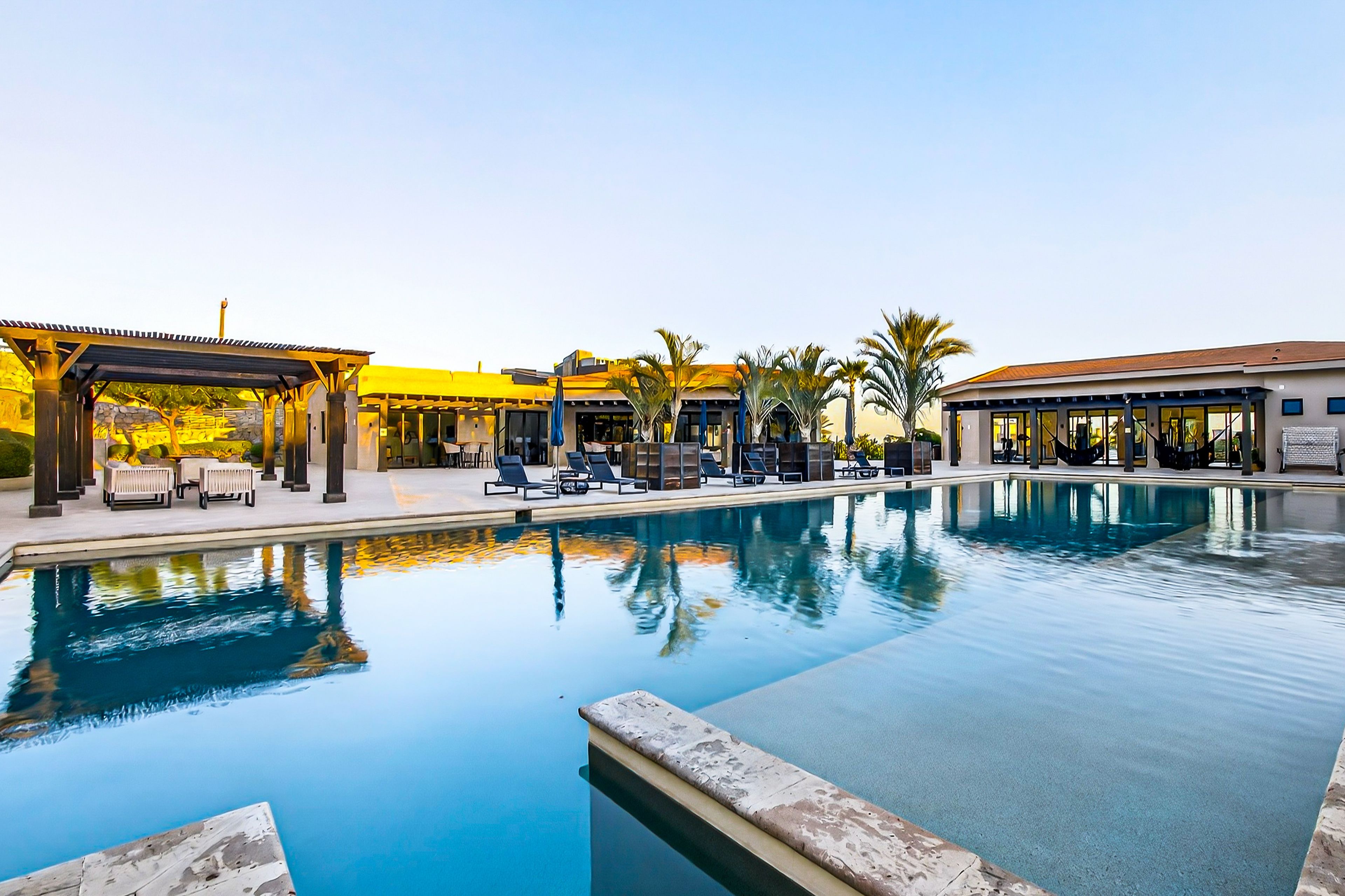 A swimming pool with lounge chairs and umbrellas, surrounded by palm trees and covered seating areas.