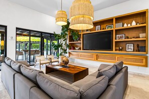 Un salon moderne avec un grand canapé d’angle, une table basse en bois et un téléviseur fixé à un mur en bois. On y trouve des étagères ornées d’objets décoratifs et une grande fenêtre donnant sur un paysage verdoyant.