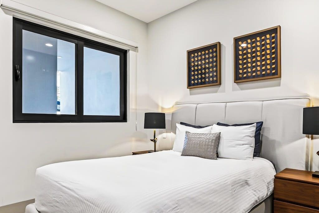 A bedroom with a large bed, two bedside tables with lamps, a window with blinds, and two wall-mounted decorative panels.