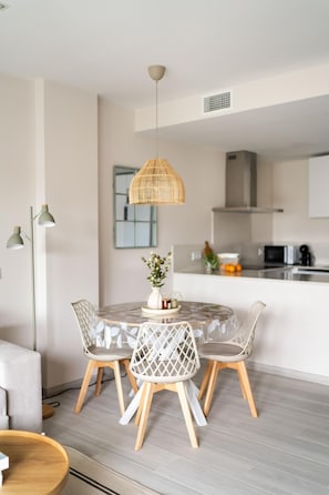 A dining area with a round table, four chairs, a hanging light fixture, and a kitchen in the background. - A lovely, brand-new apartment. 8-minute walk from the beach (AN)