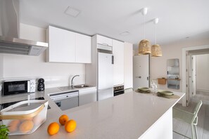 A modern kitchen with white cabinetry, a built-in oven, a microwave, and a sink. There are three oranges on the counter. - A lovely, brand-new apartment. 8-minute walk from the beach (AN)