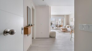 Un intérieur moderne avec un couloir menant à un espace de vie, comprenant une porte en bois, un miroir, une petite étagère et un pouf rond.
