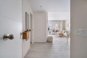 A modern interior with a hallway leading to a living area, featuring a wooden door, a mirror, a small shelf, and a round ottoman. - A lovely, brand-new apartment. 8-minute walk from the beach (AN)