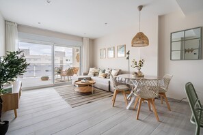 A modern living room with a sofa, coffee table, dining table, chairs, and a large window. - A lovely, brand-new apartment. 8-minute walk from the beach (AN)