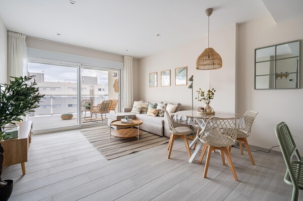 A modern living room with a sofa, coffee table, dining table, chairs, and a large window. - A lovely, brand-new apartment. 8-minute walk from the beach (AN)