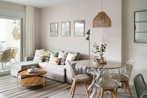 A modern living room with a sofa, a round dining table with chairs, a hanging rattan light, and framed artwork on the wall. - A lovely, brand-new apartment. 8-minute walk from the beach (AN)