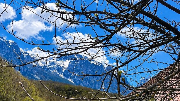 Une maison de campagne aux murs blancs et au toit rouge, entourée d’un champ verdoyant et avec une chaîne de montagnes en arrière-plan.