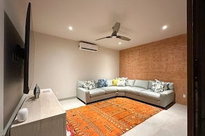 A modern living room with a grey sectional sofa, a patterned orange rug, a ceiling fan, and a flat-screen TV mounted on the wall. - Luxury House With Private Pool, Ocean View, Inviting Balcony & Elegant Charm (San José del Cabo)