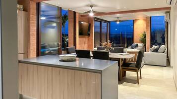 A modern kitchen with a central island, dining area with chairs, and a living room with a sofa and a TV.
