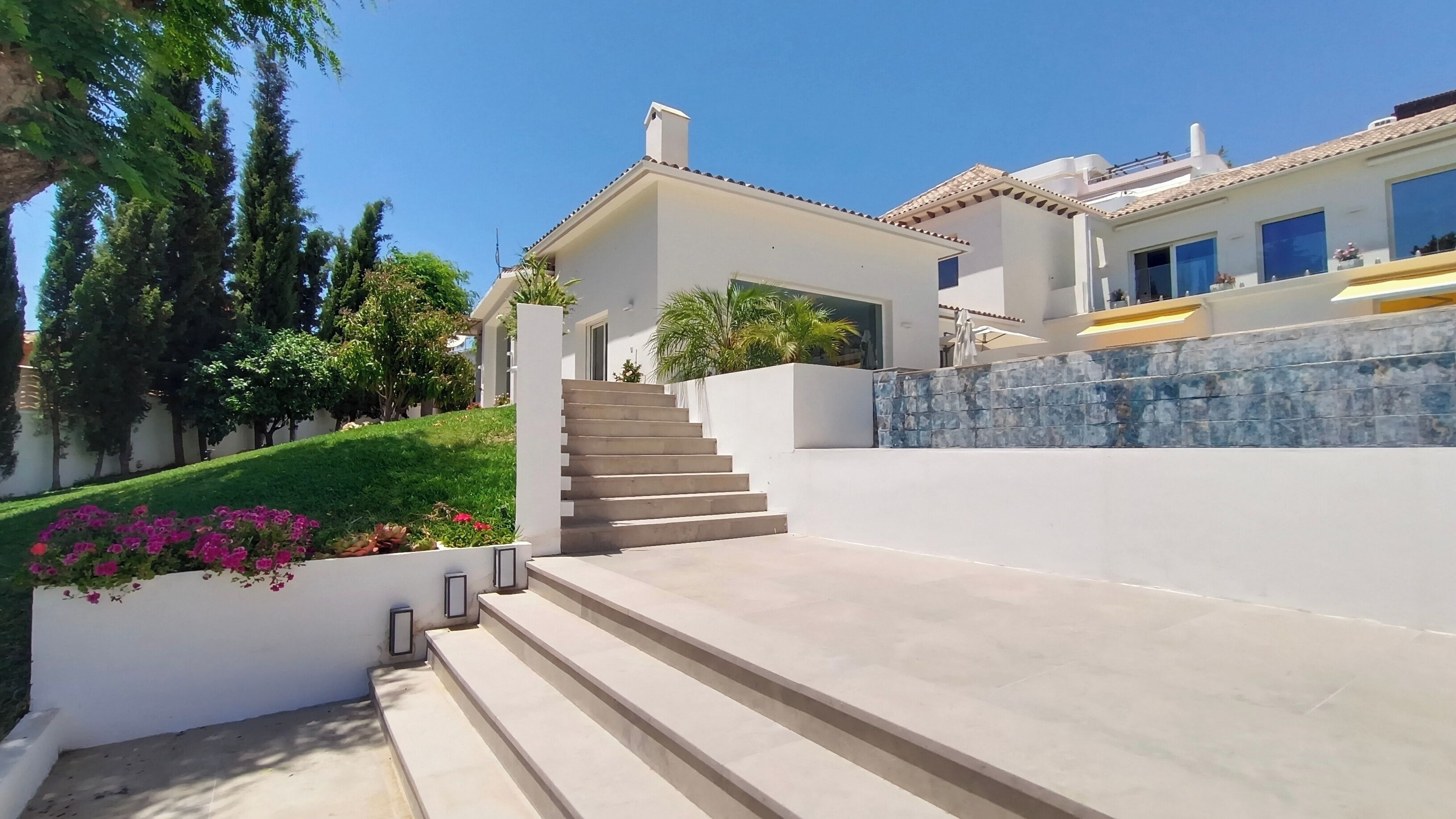A modern house with a staircase leading to a covered entrance, surrounded by greenery and a stone wall.