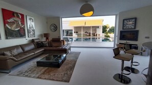 Ein modernes Wohnzimmer mit Ecksofa, Couchtisch und Essbereich mit Barhocker. Ein großes Fenster bietet Blick auf einen Pool und ein Gebäude.