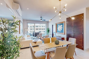 Ein modernes Wohnzimmer mit einem Glastisch, beigen Sofas, einem Deckenventilator und einem großen Fenster mit Blick auf Gebäude.