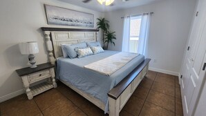 Un dormitorio con cama, mesita de noche, lámpara, ventana con cortinas, una planta y un cuadro en la pared.