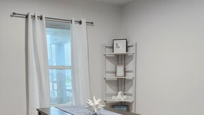 Una habitación con un escritorio de madera oscura, un estante blanco con adornos, y una ventana con cortinas blancas.