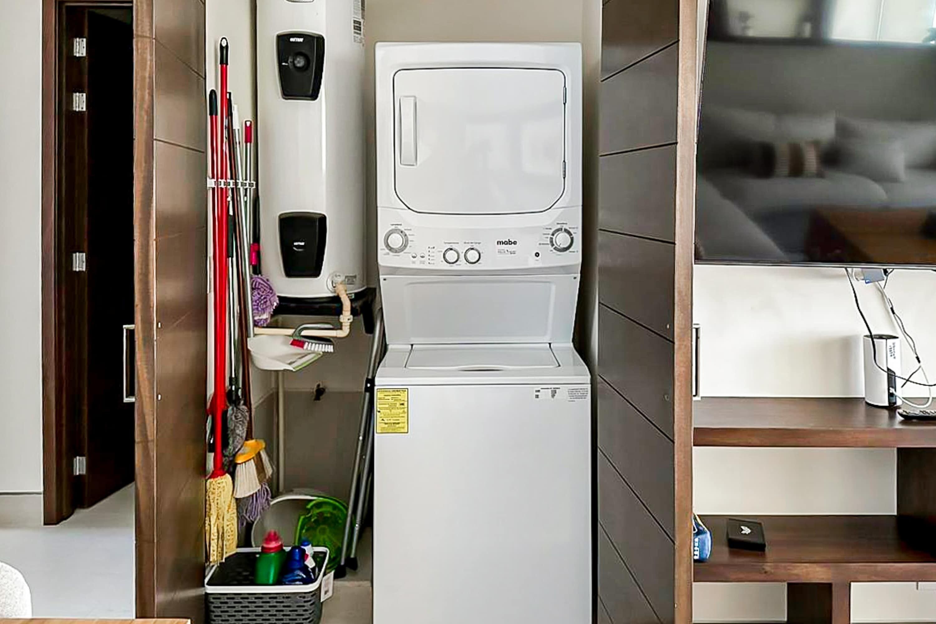 Un espace réduit dédié à la buanderie, comprenant un lave-linge et un sèche-linge, un support à balai, des produits de nettoyage et une petite étagère avec une télécommande.