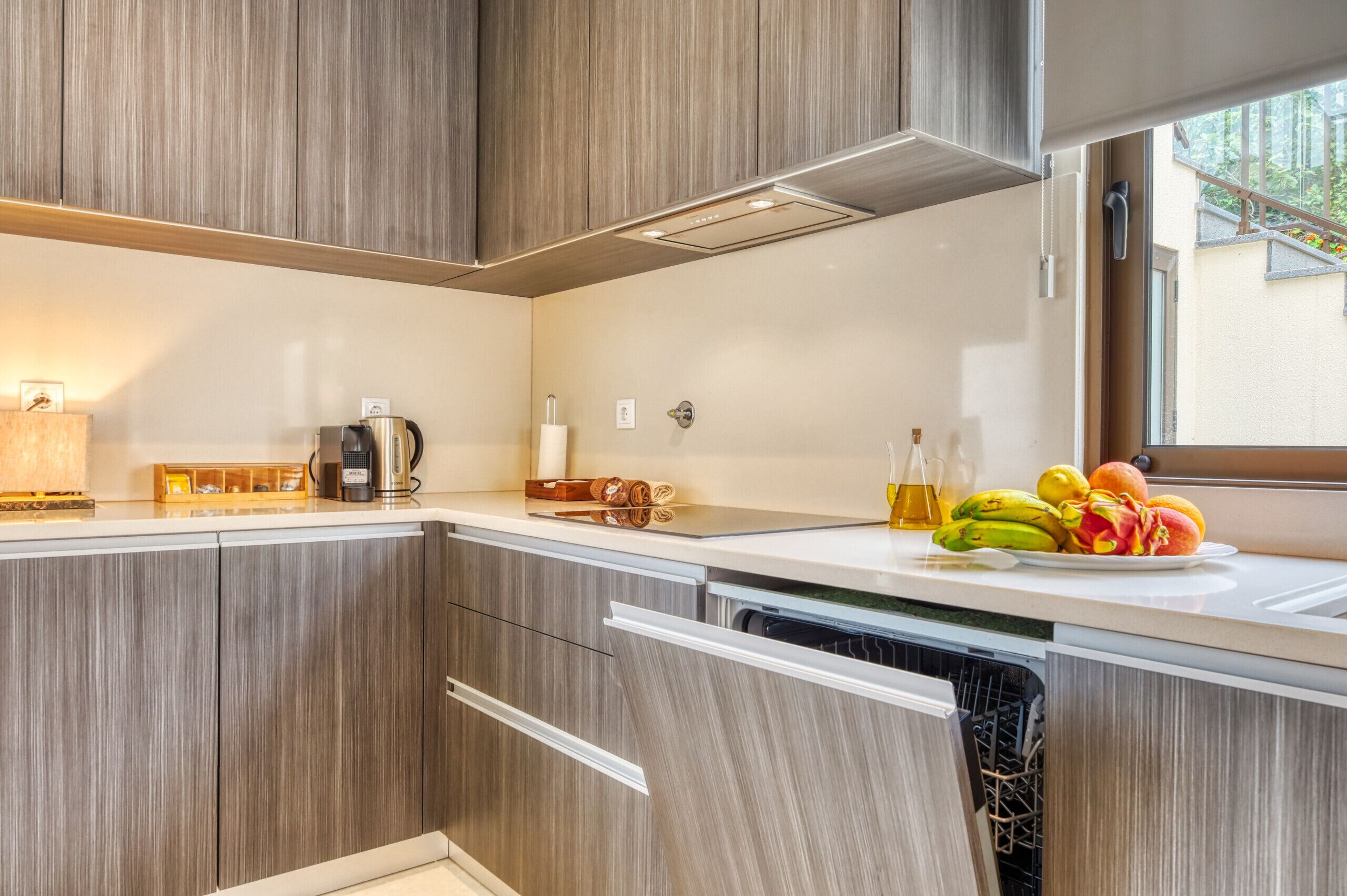 Een moderne keuken met houten kastjes, een inbouwvaatwasser, een koffiezetapparaat en een fruitschaal op het aanrecht.