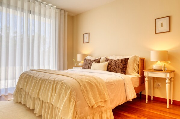 A neatly made bed with a ruffled bed skirt, two bedside lamps, and a nightstand with a lamp. - Casa da Mercês II, a Home in Madeira (São Vicente)