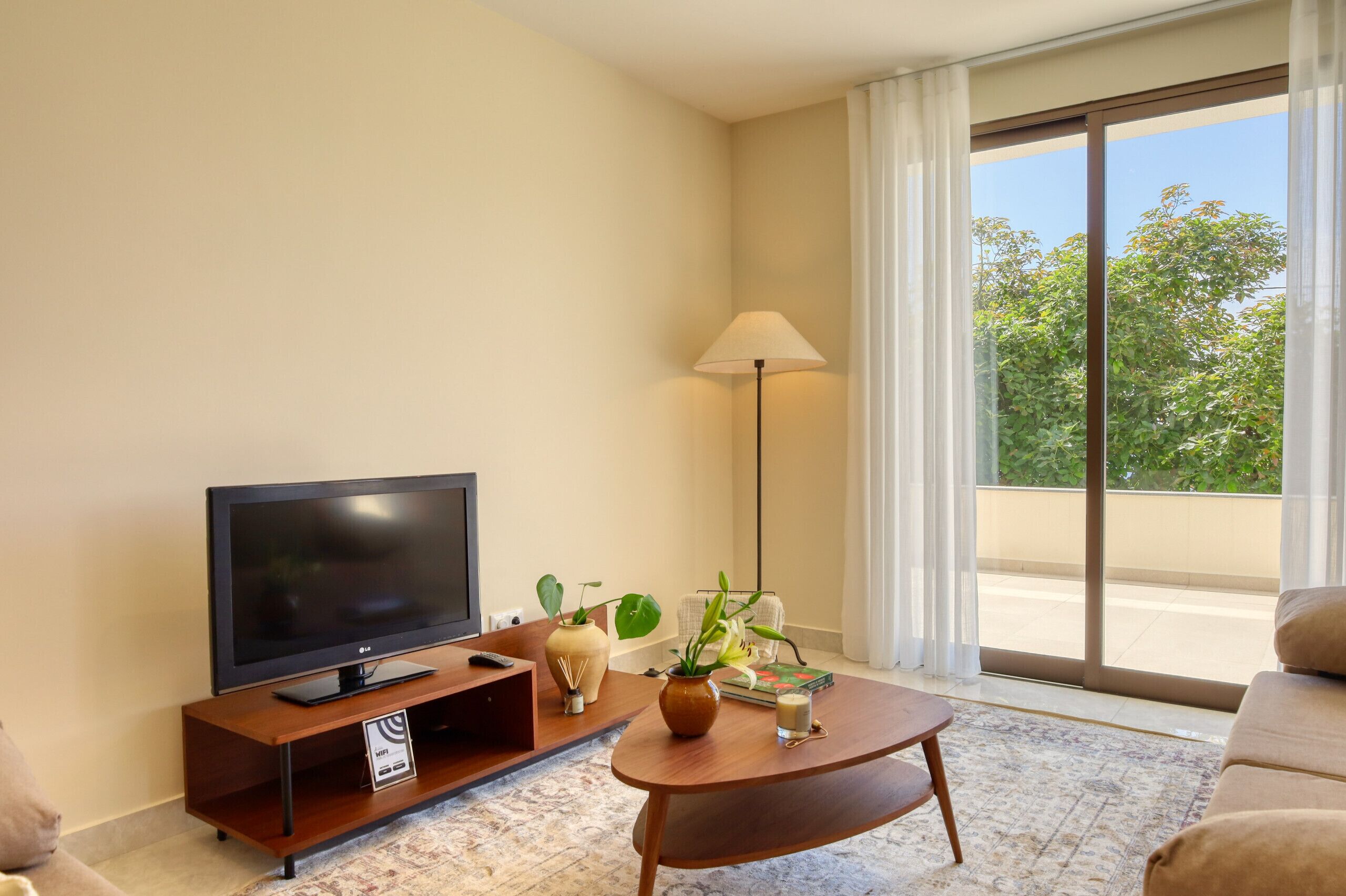 Een moderne woonkamer met een flatscreen televisie, een salontafel, een bank, een lamp en een groot raam met uitzicht op groen.