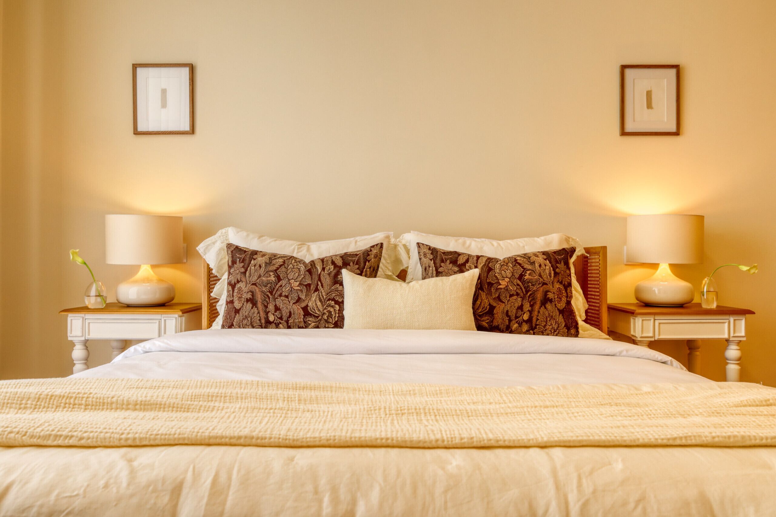 Un lit bien fait, recouvert d’une couette blanche et agrémenté de deux coussins décoratifs, avec de chaque côté des tables de chevet sur lesquelles sont posées des lampes et de petits vases.